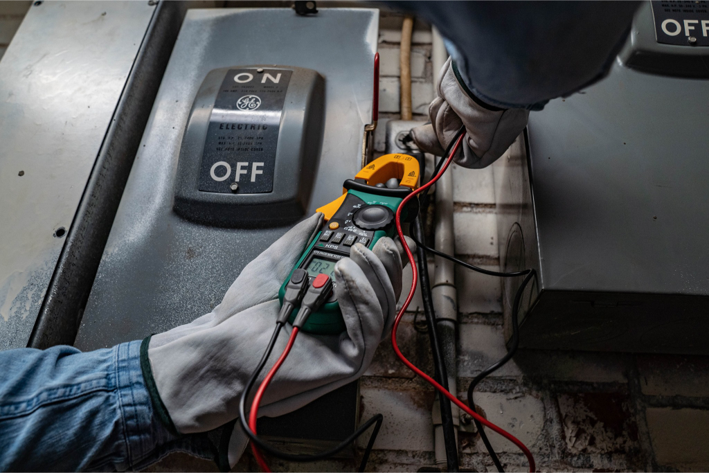 A person wearing gloves uses a multimeter to test an electrical panel beside a switch labeled "ON" and "OFF." The panel and multimeter are positioned against a brick wall.