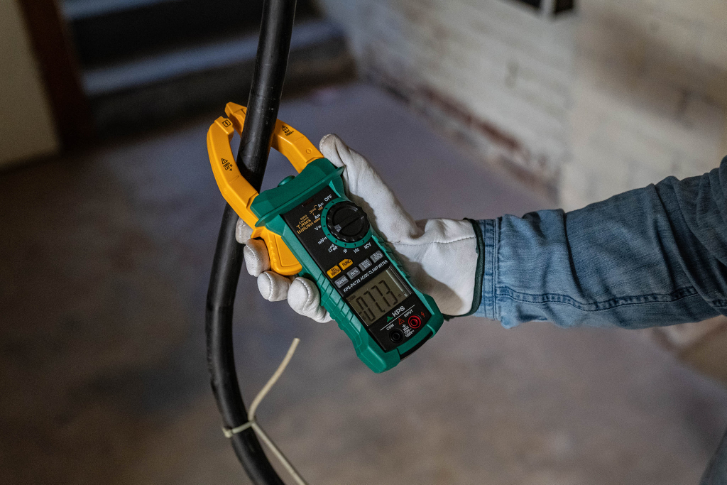 A person wearing white gloves holds a digital clamp meter with a yellow clamp around a thick black cable. The meter's display shows a reading, and the background is an indoor workspace with unfinished walls.